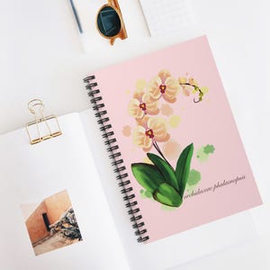 May include: A pink spiral-bound notebook with a floral illustration of a yellow orchid with red centers and green leaves. The text "orchidaceae phalaenopsis" is printed on the cover. The notebook is open on a white surface, with other stationery items.