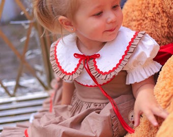May include: A young child wearing a brown dress with a white collar and red trim. The dress has a red ribbon and is paired with a large teddy bear. The child has blonde hair and is looking down.