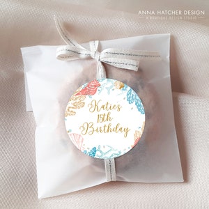 May include: A white paper bag tied with a ribbon, featuring a round sticker. The sticker has a sea-themed design with coral, seashells, and the text "Katie's 15th Birthday" in gold script.