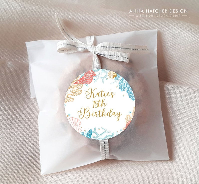 May include: A white paper bag tied with a ribbon, featuring a round sticker. The sticker has a sea-themed design with coral, seashells, and the text "Katie's 15th Birthday" in gold script.