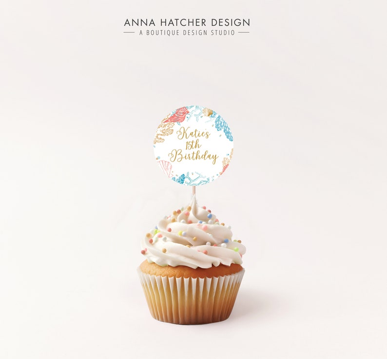 May include: A cupcake with white frosting and colorful sprinkles, topped with a round cake topper. The topper features the text "Katies 15th Birthday" in gold script, surrounded by seashell illustrations in coral, blue, and gold.
