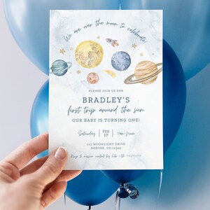 Puede incluir: Una tarjeta de invitaci&oacute;n azul y blanca con una ilustraci&oacute;n de acuarela de planetas y un cohete. El texto dice "We are over the moon to celebrate!" y "Bradley's first trip around the sun. Our baby is turning one!" La invitaci&oacute;n incluye la fecha, la hora y la ubicaci&oacute;n del evento.