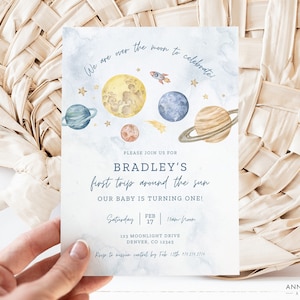 Puede incluir: Una tarjeta de invitaci&oacute;n azul y blanca con una ilustraci&oacute;n de acuarela de planetas y estrellas. El texto dice "We are over the moon to celebrate!" y "Bradley's first trip around the sun. Our baby is turning one!" La invitaci&oacute;n incluye la fecha, la hora y la direcci&oacute;n del evento.