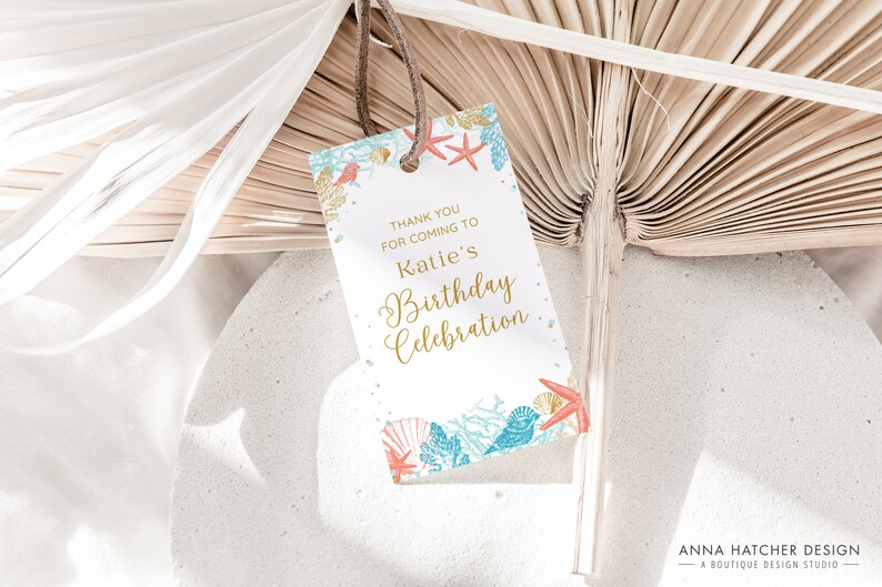 May include: A white gift tag with a beach theme, featuring text that reads "Thank you for coming to Katie's Birthday Celebration." The tag is decorated with illustrations of seashells, coral, and starfish in gold, teal, and red.