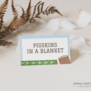 Może przedstawiać: Niebieska i biała karta z tekstem "PIGSKINS IN A BLANKET" i grafiką piłki nożnej. Karta ma zielony wz&oacute;r boiska piłkarskiego z liniami jardowymi.
