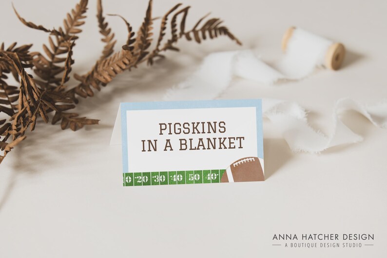 Może przedstawiać: Niebieska i biała karta z tekstem "PIGSKINS IN A BLANKET" i grafiką piłki nożnej. Karta ma zielony wz&oacute;r boiska piłkarskiego z liniami jardowymi.