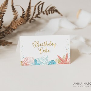 May include: A white folded card with the words "Birthday Cake" in gold script. The card features illustrations of seashells, coral, and a starfish in shades of coral, blue, and gold. The card is designed for a birthday celebration.