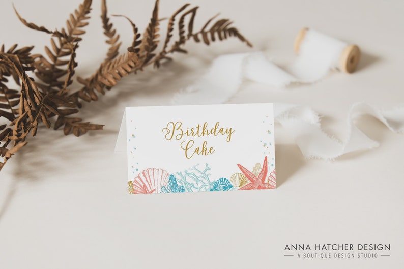 May include: A white folded card with the words "Birthday Cake" in gold script. The card features illustrations of seashells, coral, and a starfish in shades of coral, blue, and gold. The card is designed for a birthday celebration.