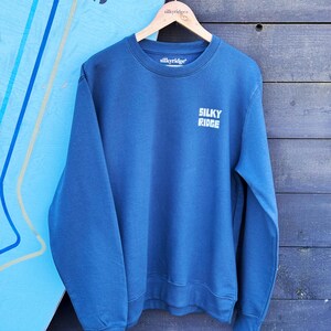 Könnte beinhalten: Ein blaues Sweatshirt mit dem Text "SILKY RIDGE" in weißen Blockbuchstaben auf der Vorderseite. Das Sweatshirt hängt an einem Holzbügel. Ein hellblaues Surfbrett ist im Hintergrund.