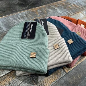 Puede incluir: Cinco gorros de punto de diferentes colores, incluyendo azul verdoso, beige, rosa y verde. Cada gorro tiene un parche de madera con un diseño geométrico.