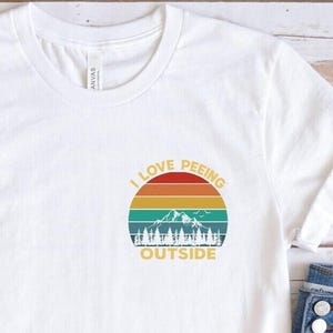 May include: White t-shirt with a colorful graphic that reads "I LOVE PEEING OUTSIDE" in a circular design with a mountain and tree silhouette. The image also includes denim shorts, brown sandals, and sunglasses.