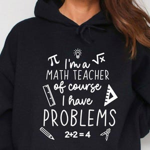 May include: Black hooded sweatshirt with white text that reads "I'm a math teacher of course I have PROBLEMS". The design includes mathematical symbols like pi, a square root, and a ruler. The sweatshirt is paired with denim shorts.