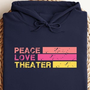 May include: A navy blue hoodie with the words "PEACE LOVE THEATER" in a distressed, colorful design. The text is stacked with each word on a separate horizontal bar of color: peach, pink, and yellow. The hoodie is folded on a woven surface.