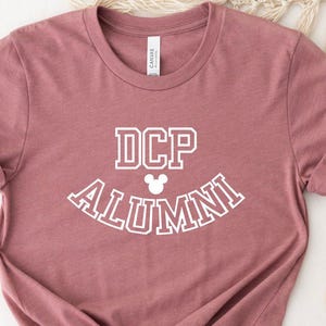 May include: A mauve t-shirt with white text that reads "DCP ALUMNI" with a white Mickey Mouse head in the middle of the text.