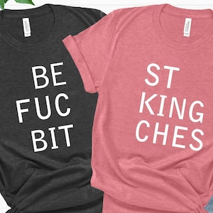 May include: Two short-sleeved t-shirts, one dark gray and one mauve, with white text. The dark gray shirt reads "BE FUC BIT" and the mauve shirt reads "ST KING CHES". The shirts are knotted at the bottom.