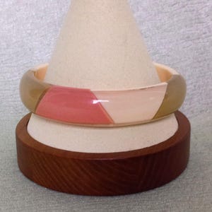 May include: A colorful bangle bracelet with a geometric design. The bracelet features sections of peach, tan, and cream colors. The bracelet is displayed on a white cone-shaped stand, which sits on a wooden base.