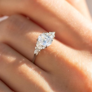 May include: A silver engagement ring with a large, round, clear center stone and smaller accent stones. The ring is set on a delicate band and is held up by a hand.
