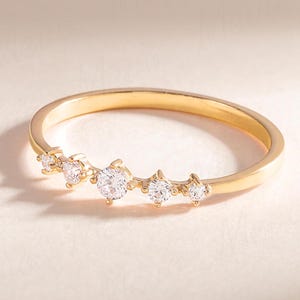 Może przedstawiać: Złoty pierścionek z rzędem małych, przezroczystych kamieni ułożonych w zakrzywioną linię.