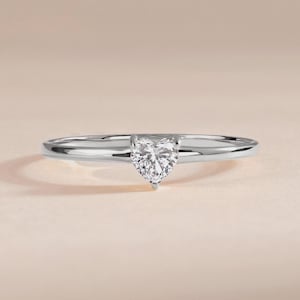 May include: A silver ring with a heart-shaped diamond. The ring has a slender, polished band, and the diamond is held in place by prongs. The ring is presented against a soft, neutral backdrop, emphasising its elegant design.
