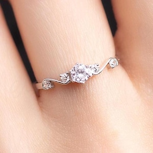 Könnte beinhalten: Ein zarter Silberring mit drei klaren, runden Edelsteinen, die in einem wirbelnden Design gefasst sind. Der Ring hat ein schlankes Band und kleine Akzentsteine auf jeder Seite der zentralen Steine. Der Ring wird an einem Finger gezeigt.