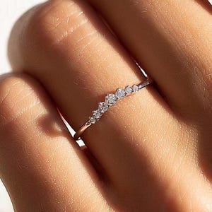 Può includere: Un anello sottile e delicato con una fila di piccoli diamanti scintillanti disposti in un design curvo. L'anello è di un colore metallico chiaro, forse oro o argento, e viene indossato su un dito. L'attenzione è sull'elegante semplicità dell'anello e sulla brillantezza delle gemme.