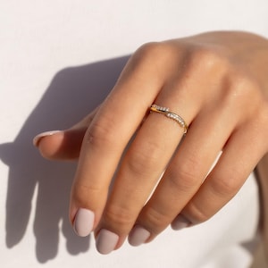 Pode incluir: Um anel dourado com um design curvo, adornado com pequenas pedras preciosas transparentes. O anel &eacute; usado em uma m&atilde;o com esmalte rosa claro, em um fundo branco.