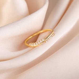 Pode incluir: Um anel dourado com um design delicado. O anel apresenta uma banda que se curva e se cruza, adornada com pequenas pedras preciosas transparentes. O anel est&aacute; sobre um tecido macio de cor p&ecirc;ssego.