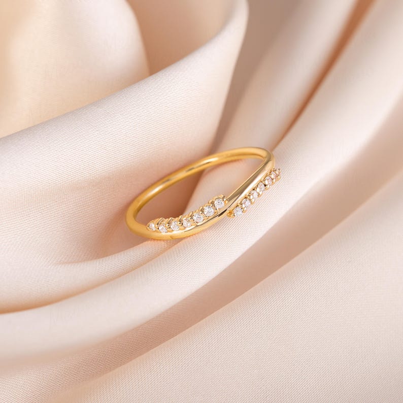 Pode incluir: Um anel dourado com um design delicado. O anel apresenta uma banda que se curva e se cruza, adornada com pequenas pedras preciosas transparentes. O anel est&aacute; sobre um tecido macio de cor p&ecirc;ssego.