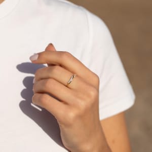 Pode incluir: Um anel de ouro com pequenas pedras preciosas transparentes dispostas em um design curvo. O anel &eacute; usado em uma m&atilde;o, com uma camiseta branca ao fundo. O design delicado do anel e o brilho das pedras preciosas s&atilde;o destacados.