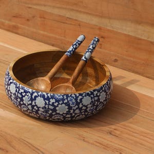 Puede incluir: Un cuenco de ensalada de madera con un diseño floral azul y blanco alrededor del borde. Dos cucharas de madera con mangos estampados en azul y blanco descansan dentro del cuenco. El cuenco y las cucharas están sobre una superficie de madera.