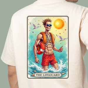 以下が含まれることがあります： クリーム色のTシャツの背面に、サングラス、赤い水着、オレンジ色のライフジャケットを着用した骸骨のライフガードのグラフィックが描かれています。イラストには明るい太陽、カモメ、そして下部に「THE LIFEGUARD」という文字が含まれています。