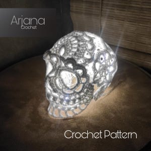 Könnte beinhalten: Eine weiße, gehäkelte Totenkopflampe mit einem Licht im Inneren, das einen warmen Schein erzeugt. Der Totenkopf ist mit komplizierten Häkelmustern verziert. Der Text "Crochet Pattern" ist unterhalb des Totenkopfs sichtbar.