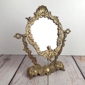 Peut inclure: Miroir de courtoisie doré orné, avec un cadre ovale. Le miroir présente des sculptures en relief détaillées de chérubins, de coquillages et de motifs floraux. Le miroir est soutenu par une base décorative et possède deux poignées latérales.
