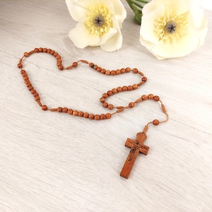 Könnte beinhalten: Ein hölzerner Rosenkranz mit braunen Perlen und einem Kruzifix. Die Perlen sind mit einer hellbraunen Schnur verbunden. Das Kruzifix zeigt ein geschnitztes Bild von Jesus. Der Rosenkranz wird auf einer hellen Holzoberfläche präsentiert.