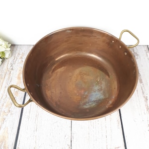 Peut inclure: Une marmite de cuisine en cuivre vintage avec deux poignées en laiton. La marmite est de forme ronde et présente un aspect vieilli et terni. Les poignées sont fixées sur les côtés de la marmite. La marmite est posée sur une surface en bois blanc.