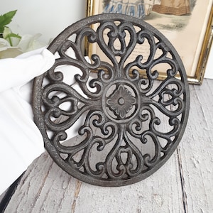 Peut inclure: Un dessous de plat circulaire en fonte gris foncé présentant un motif ajouré complexe de volutes symétriques et un motif floral central. La surface métallique a une finition texturée, et le design se compose de lignes courbes répétitives et d'éléments en forme de cœur disposés en motif radial autour du point central.