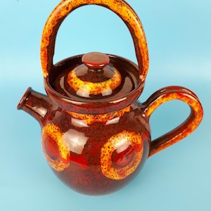 Peut inclure: Une théière en céramique brune avec un glaçage jaune et orange. La théière a un corps rond avec une poignée circulaire et un couvercle avec un bouton.