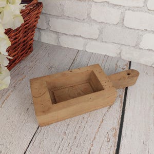 May include: A wooden mold for making soap or other treats. The mold has a rectangular shape with a carved design on the inside. It has a handle on one side.