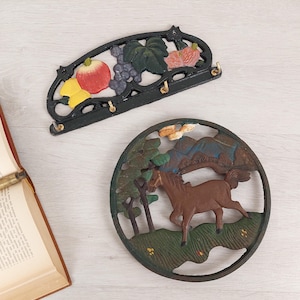 Può includere: Due pezzi di arredamento da parete in ghisa. Uno è un portachiavi nero con un design a vite e frutta. L'altro è un pezzo rotondo in ghisa nera con un cavallo marrone che corre attraverso una scena di foresta verde.