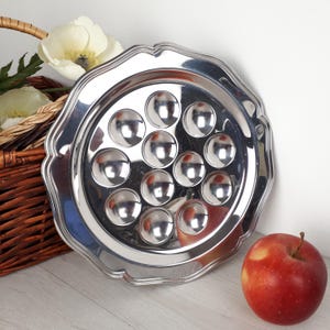 Peut inclure: Un plat à escargots argenté brillant avec douze cavités rondes. Le plateau à bord festonné est à côté d'une pomme rouge et d'un panier en osier avec des fleurs blanches.