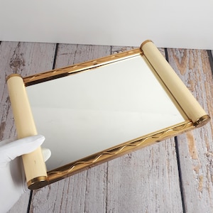 Peut inclure: Miroir de courtoisie rectangulaire avec un cadre doré et des poignées cylindriques crème. Le cadre présente un motif ondulé. Le miroir est tenu par une main gantée sur une surface en bois vieilli.