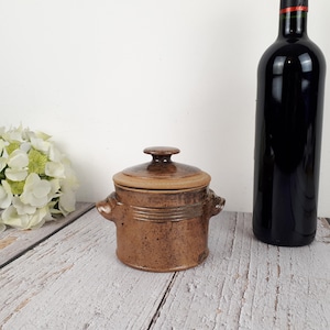 以下が含まれることがあります： ふたと2つの取っ手がついた茶色の陶器の壺。壺は斑点のある釉薬がかかっており、木製の表面の上に置かれています