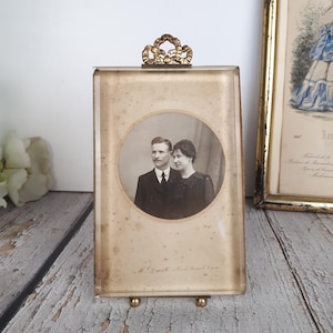 Peut inclure: Un presse-papier ou cadre rectangulaire en verre contenant une photographie circulaire sépia d'un couple. Le verre est surmonté d'un nœud décoratif en métal doré et repose sur deux petits pieds en métal doré. Le verre présente des signes d'usure.