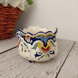 Peut inclure: Petit pot de fleurs en céramique blanc cassé, à bord festonné et trois petits pieds. Il est orné d'un motif floral aux couleurs vives : bleu, rouge, jaune et vert. Le bord est bordé d'un liseré bleu.
