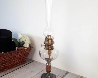 May include: A tall, antique oil lamp with a clear glass chimney, a round glass reservoir, and a brass burner. The lamp has a wooden base and a metal base with the word "LUX" inscribed on it.