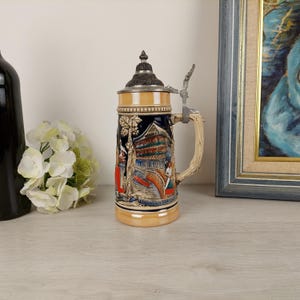 Peut inclure: Une chope à bière en céramique avec une scène détaillée d'un village allemand, comprenant un pont, un bâtiment avec une enseigne indiquant "Gasthof der Traube" et des personnes en costumes traditionnels. La chope est munie d'un couvercle avec une poignée en métal.