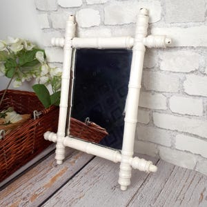 Peut inclure: Un miroir encadré de style bambou blanc. Le miroir rectangulaire a une surface sombre et réfléchissante. Le cadre a une finition vieillie. Le miroir est posé sur une surface en bois. Le cadre imite le bambou.