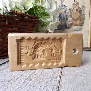 Peut inclure: Un moule à beurre en bois clair avec un motif sculpté détaillé. Le moule rectangulaire présente un motif central de coq et de blé, entouré d'une bordure décorative. Un petit trou est présent à une extrémité.