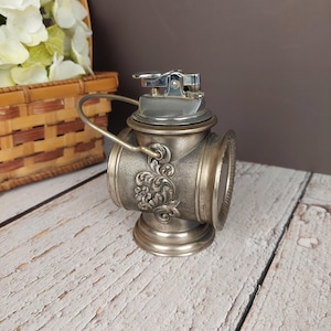 May include: Silver-toned table lighter shaped like a vintage lantern with a decorative floral design. The lighter has a hinged lid and a small handle.