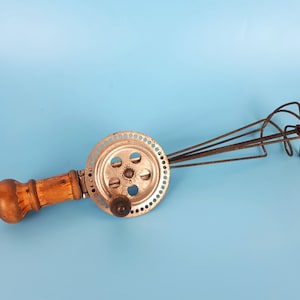 May include: A vintage hand-crank egg beater with a wooden handle and a metal whisk. The whisk has multiple wires that are attached to a circular metal gear.
